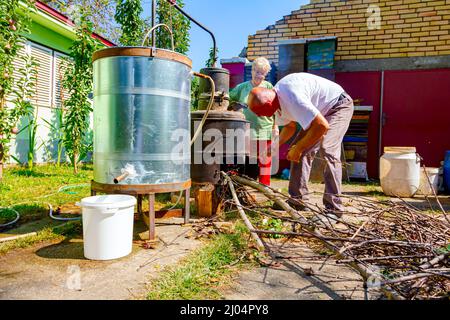 La femme tourne manuellement le levier pour mélanger le Marc de fruits dans la chaudière de la distillerie faite maison en cuivre, faisant brandy et l'homme jette des branches sèches dans le Banque D'Images