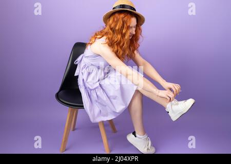 Femme à tête rouge pleine longueur, jeune femme en chapeau de paille et en poussette assise sur la chaise et nouant des lacets sur les éternuements blancs. Banque D'Images