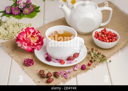 Tisane à la rosehip, camomille et limbes dans une tasse blanche sur une table blanche en bois avec des fleurs. Banque D'Images