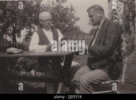 photo vintage originale d'un grand-père et petit-fils qui sont assis autour d'une table et carte à jouer (carte à jouer allemande) des années 1930 à l'heure d'été. / homme vintage fumant une cigarette Banque D'Images