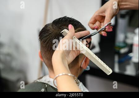 Un adolescent dans un salon de beauté obtient une coupe de cheveux, un coiffeur coupe les cheveux d'un garçon adolescent, une coupe de cheveux avec des ciseaux et un peigne. Banque D'Images