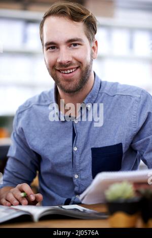 Le design est sa passion. Portrait d'un beau jeune homme à travers un magazine au bureau. Banque D'Images