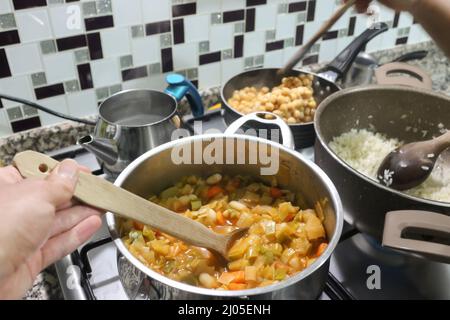 Cuisson de plat de légumes et de riz sur la cuisinière dans la cuisine. Main tenant une cuillère en bois. Carotte, poireau, haricots, chou, pâte de tomate. Concept de cuisine végétalienne Banque D'Images