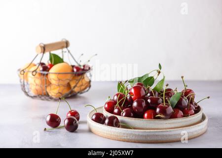 Cerises fraîches dans deux assiettes en céramique légère et abricots dans un panier en fer osier sur un fond gris et blanc. Banque D'Images
