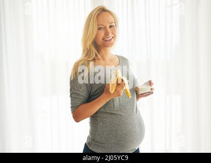 Maman en bonne santé, bébé en bonne santé. Portrait d'une femme enceinte heureuse mangeant une banane et buvant un verre de lait à la maison. Banque D'Images