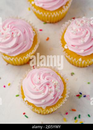 Vue de dessus. Petits gâteaux à la crème rose fouettée sur fond blanc et confettis colorés. Composition festive. Anniversaire, fête, vacances en famille Banque D'Images