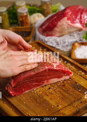 Gros plan. Les mains du chef frottent les épices sur un gros morceau de viande sur une planche à découper en bois. Plats de cuisson à base de viande, recettes de porc, bœuf. Restaurant, c Banque D'Images