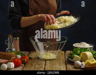 Le chef met du fromage râpé dans un bol en verre. Les légumes, les œufs, les herbes, les épices se trouvent sur une table en bois. Préparation de salades de légumes, pizzas, pâtes. H Banque D'Images