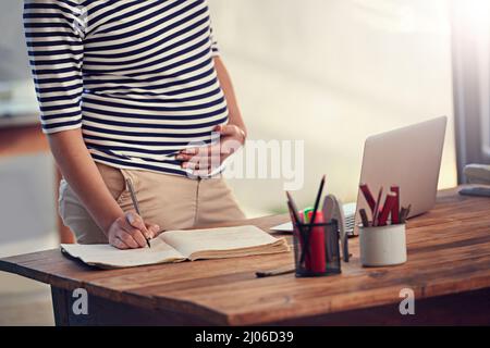 S'assurer qu'elle sait exactement ce qui doit être fait avant le congé de maternité. Prise de vue rognée d'une jeune femme enceinte qui écrit dans son journal tout en étant debout Banque D'Images