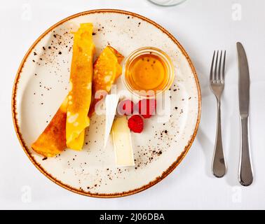 Crêpes préparées sur une assiette avec framboises, tranches de fromage et miel, sur fond blanc. Banque D'Images