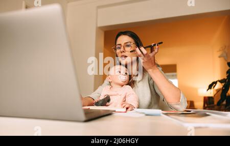 Une nouvelle maman qui parle lors d'un appel téléphonique dans son bureau à domicile. Maman multitâche travaillant sur un nouveau projet créatif à son bureau. Femme de conception d'intérieur de transport Banque D'Images