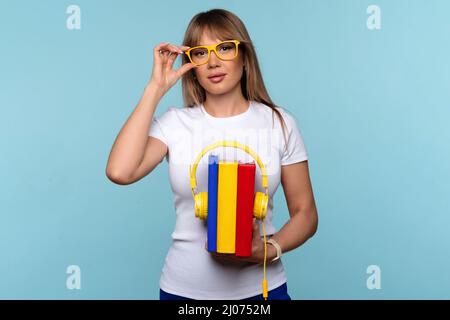 Femme en t-shirt blanc et en lunettes tenant des livres et un casque. Les casques jaunes ont placé trois livres en arrière. Livres audio. Écoutez et apprenez. Formation en ligne et e-learning. Enregistrement de podcast Banque D'Images