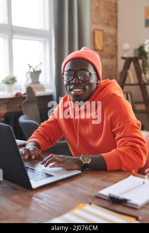 Portrait vertical d'un homme afro-américain joyeux portant une tenue orange vif assis au bureau à la maison surfant sur Internet sur un ordinateur portable souriant à l'appareil photo Banque D'Images