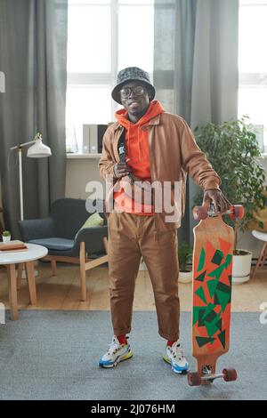 Portrait vertical complet d'un jeune homme élégant debout dans le salon avec planche à roulettes prête à sortir en souriant à l'appareil photo Banque D'Images