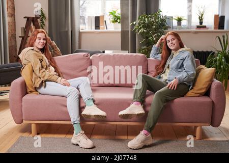 Photo large de deux belles jeunes sœurs caucasiennes jumelles avec de longs cheveux rouges portant des tenues décontractées assis sur un canapé dans le salon Banque D'Images