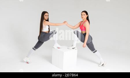 Deux femmes de fitness donnent poignée de main tout en se tenant sur le cube en studio. Concept de travail d'équipe sportif Banque D'Images