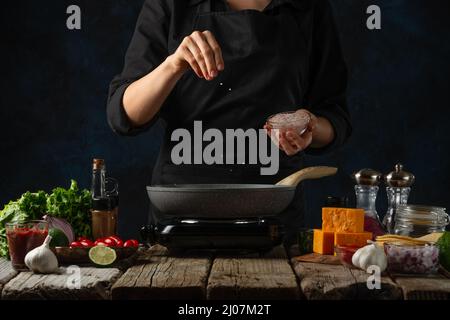 Chef professionnel en uniforme noir verse du sel de mer dans une poêle avec garniture de viande. Arrière-plan de la cuisine traditionnelle mexicaine tacos sur fond bleu foncé Banque D'Images
