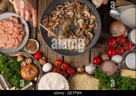 Les mains du chef remuent les champignons dans la poêle pour préparer le remplissage sur une table en bois avec une variété d'ingrédients de fond. Concept du processus de cuisson. Banque D'Images