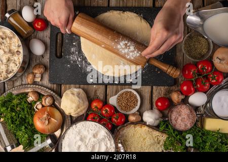 Le chef fait rouler la pâte avec une broche sur une table en bois avec une variété d'ingrédients de fond. Concept du processus de cuisson. Arrière-plan de la cuisson Banque D'Images