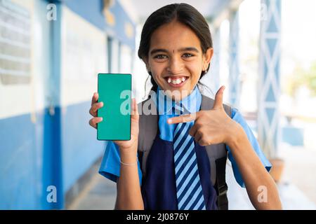 Sourire fille d'école enfant en uniforme montrant vert écran téléphone mobile en pointant plus fin tout en regardant la caméra dans le couloir - concept de l'éducation Banque D'Images