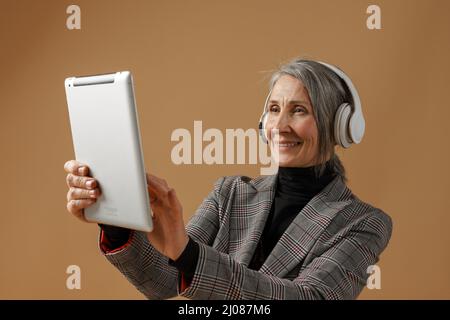 Femme grise âgée dans un casque souriant et utilisant un ordinateur isolé sur fond beige Banque D'Images