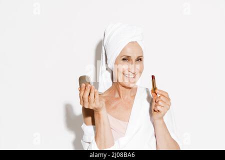Femme âgée portant une serviette souriante tout en appliquant un rouge à lèvres isolé sur un mur blanc Banque D'Images