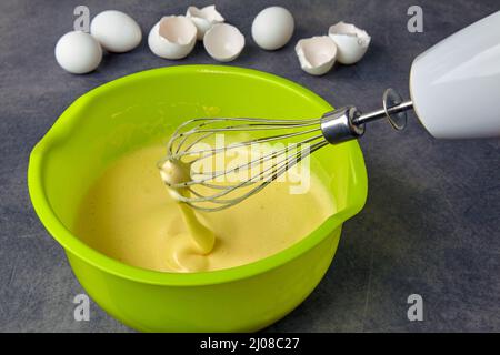 Battez les œufs avec du sucre dans un bol vert et un fouet contre le fond d'une coquille d'œuf sur une table en béton foncé Banque D'Images