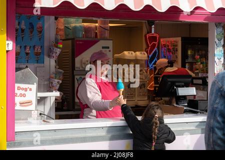 Un vendeur de crème glacée à un kiosque en bord de mer passe une glace bleue dans un cône à une jeune fille avec une longue tresse. Banque D'Images