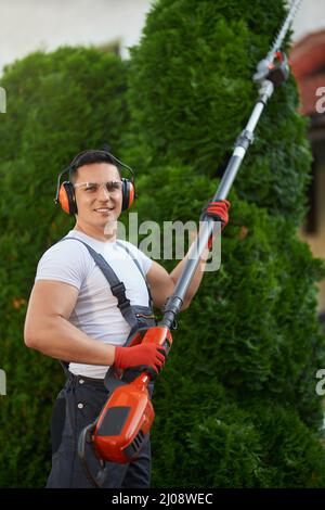 Homme caucasien positif avec taille-haie à essence dans les mains debout sur la cour arrière et regardant la caméra. Jardinier compétent pour tondre les plantes à l'air frais. Banque D'Images