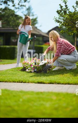 Femme caucasienne aux cheveux blonds mettant les fleurs de semis dans le sol tandis que la jeune fille les arrosant derrière. Mère et fille prenant soin des plantes sur la cour arrière. Banque D'Images