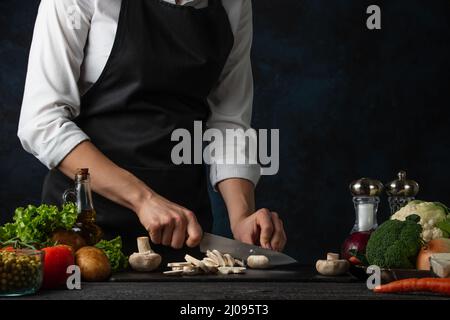 Le chef professionnel en tablier noir coupe avec des champignons au couteau sur le tableau haché pour cuire de la soupe sur fond bleu foncé. Arrière-plan de préparation m Banque D'Images