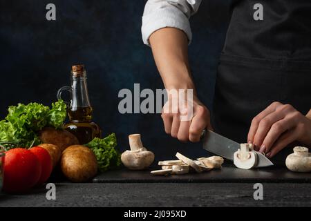 Vue macro du chef en tablier noir avec des couteaux de champignons sur le tableau haché pour cuire la soupe sur fond bleu foncé. Arrière-plan de la préparation Banque D'Images