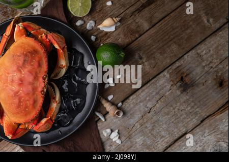 Vue de dessus d'une assiette ronde noire avec crabe cuit servi avec des glaçons, des coquillages et de la chaux sur fond de bois. Repas délicieux. Concept de fruits de mer. Espace Banque D'Images