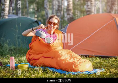 Une femme dans un sac de couchage boit du thé sur fond de nombreuses tentes dans un camping tôt le matin Banque D'Images