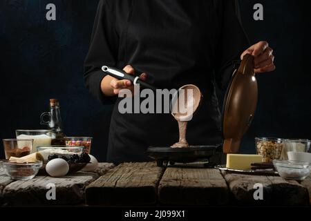 Gros plan du chef verse de la pâte dans une machine à gaufres. L'arrière-plan de la cuisine est composé de gaufres sur une table en bois rustique avec des ingrédients sur fond bleu foncé. Banque D'Images