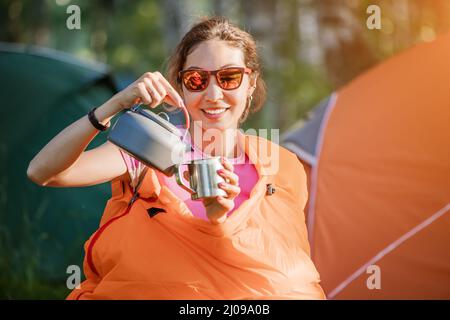 Une femme dans un sac de couchage boit du thé sur fond de nombreuses tentes dans un camping tôt le matin Banque D'Images