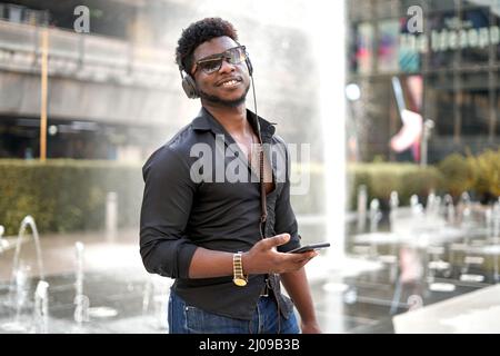 Homme noir souriant à l'appareil photo tout en écoutant de la musique avec le mobile en extérieur Banque D'Images