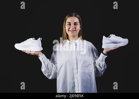 Une femme aux cheveux justes dans une chemise blanche sur fond noir tient des baskets blanches en cuir véritable dans ses mains Banque D'Images