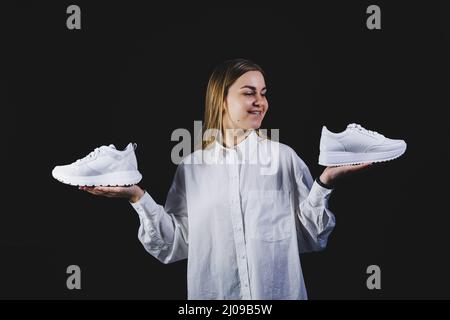 Une femme aux cheveux justes dans une chemise blanche sur fond noir tient des baskets blanches en cuir véritable dans ses mains Banque D'Images