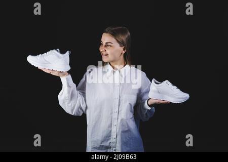 Une femme aux cheveux justes dans une chemise blanche sur fond noir tient des baskets blanches en cuir véritable dans ses mains Banque D'Images