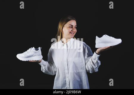 Une femme aux cheveux justes dans une chemise blanche sur fond noir tient des baskets blanches en cuir véritable dans ses mains Banque D'Images