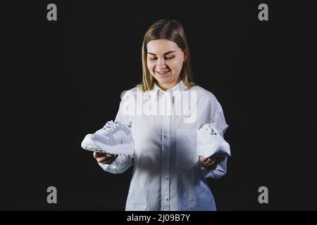 Une femme aux cheveux justes dans une chemise blanche sur fond noir tient des baskets blanches en cuir véritable dans ses mains Banque D'Images