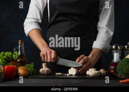 Vue macro des coupes de chef avec des couteaux de champignons sur le tableau haché pour cuire de la soupe sur fond bleu foncé. Coulisses de la préparation du repas. Plat sain Banque D'Images