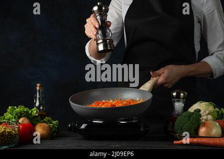 Le chef professionnel verse du poivre dans une poêle avec des légumes friture pour cuire de la soupe sur fond bleu foncé. Coulisses de la préparation du repas. Plat sain. Pour Banque D'Images