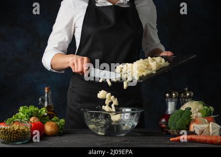 Le chef professionnel verse des pommes de terre hachées dans un bol en verre pour cuire de la soupe sur fond bleu foncé. Coulisses de la préparation du repas. Plat sain. Cuisine con Banque D'Images