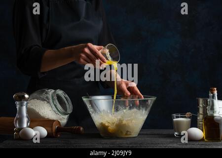 Le chef professionnel verse du jus d'orange dans le bol en verre pour préparer la pâte. Ingrédients pour cuisiner en arrière-plan. Fond bleu foncé spac Banque D'Images