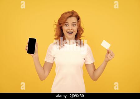 une jeune femme excitée a isolé un fond surjaune en utilisant un téléphone portable tenant une carte de crédit. Banque D'Images