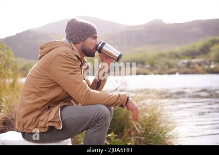 Profiter du paysage. Photo d'un beau homme assis près d'un lac buvant à partir d'un thermos. Banque D'Images