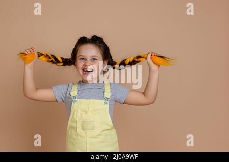 Jeune fille rayonnante et excitée avec des tresses de kanekalon de jaune, faisant grimace tirant des queues de porc avec les mains regardant la caméra sourire porter Banque D'Images