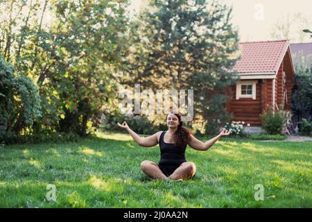 Jeune femme sur-pondérée faisant du yoga assis dans la position de lotus à l'extérieur de l'arrière-cour de la maison avec des arbres et maison en bois en arrière-plan. Corps positif Banque D'Images